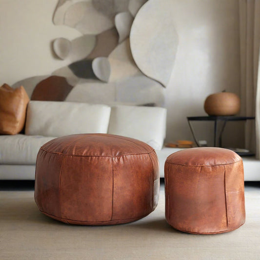 Two Moroccan leather poufs, one large and one small, both handcrafted with natural leather and detailed stitching, arranged together in a stylish living room with modern furnishings.