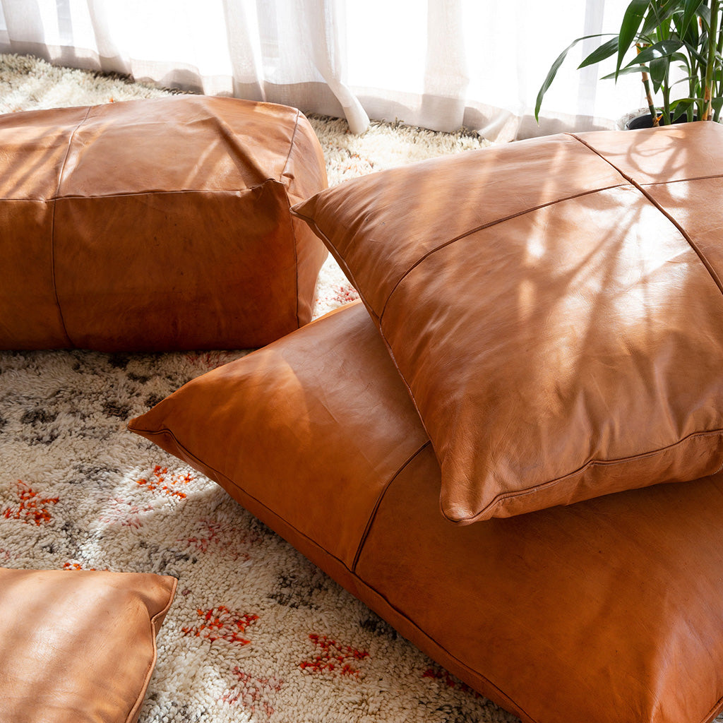 Handcrafted Moroccan leather pillows in tan color, showcasing square and rectangular shapes, placed on a textured rug in a cozy living room setting.