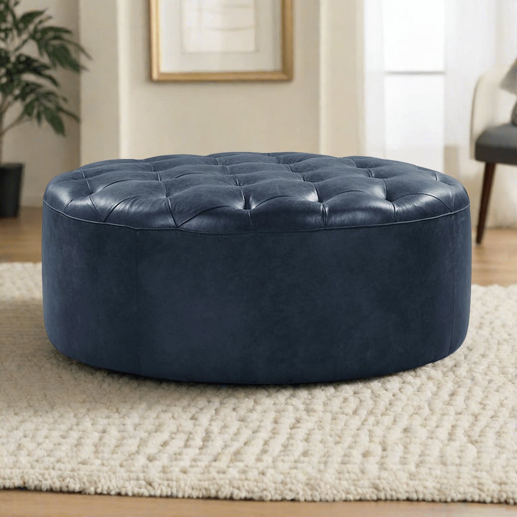 A round, tufted leather ottoman in a dark navy blue color, set on a light beige rug in a bright living room featuring a framed picture on the wall and a modern chair in the background.