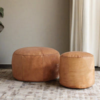 A pair of handcrafted Moroccan leather poufs, one larger and one smaller, both featuring intricate hand-stitching, placed on a patterned rug in a neutral-toned room with elegant decor.