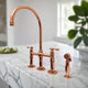 Polished copper bridge kitchen faucet with side sprayer installed on white marble countertop in a bright modern kitchen
