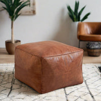 Tan leather cube ottoman placed in a cozy room with a yellow chair and a side table in the background.